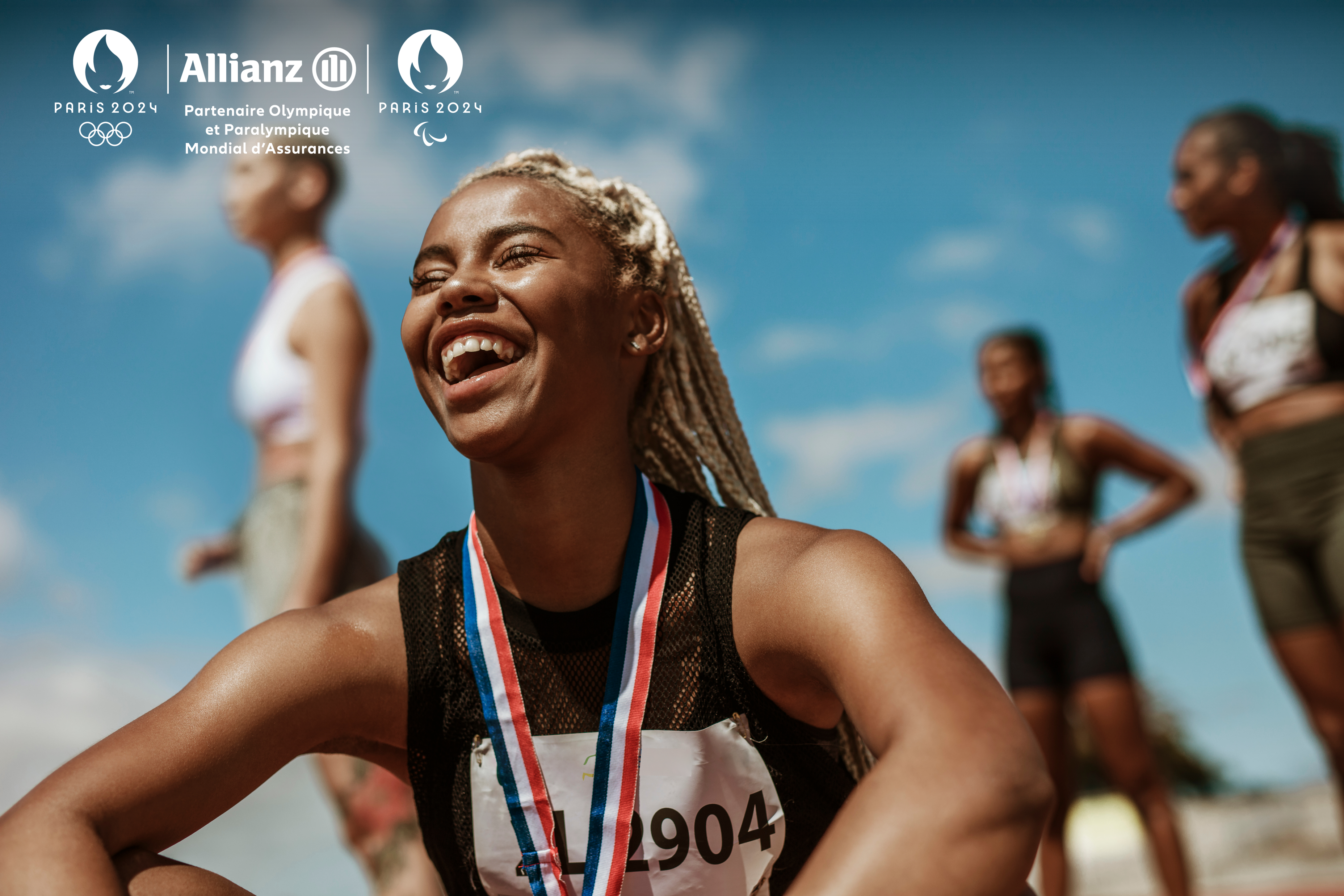 Black female athlete sitting with arms over knees laughing. She had a red white and blue ribbon around her neck and three other  athletes are standing behind her (blurred).