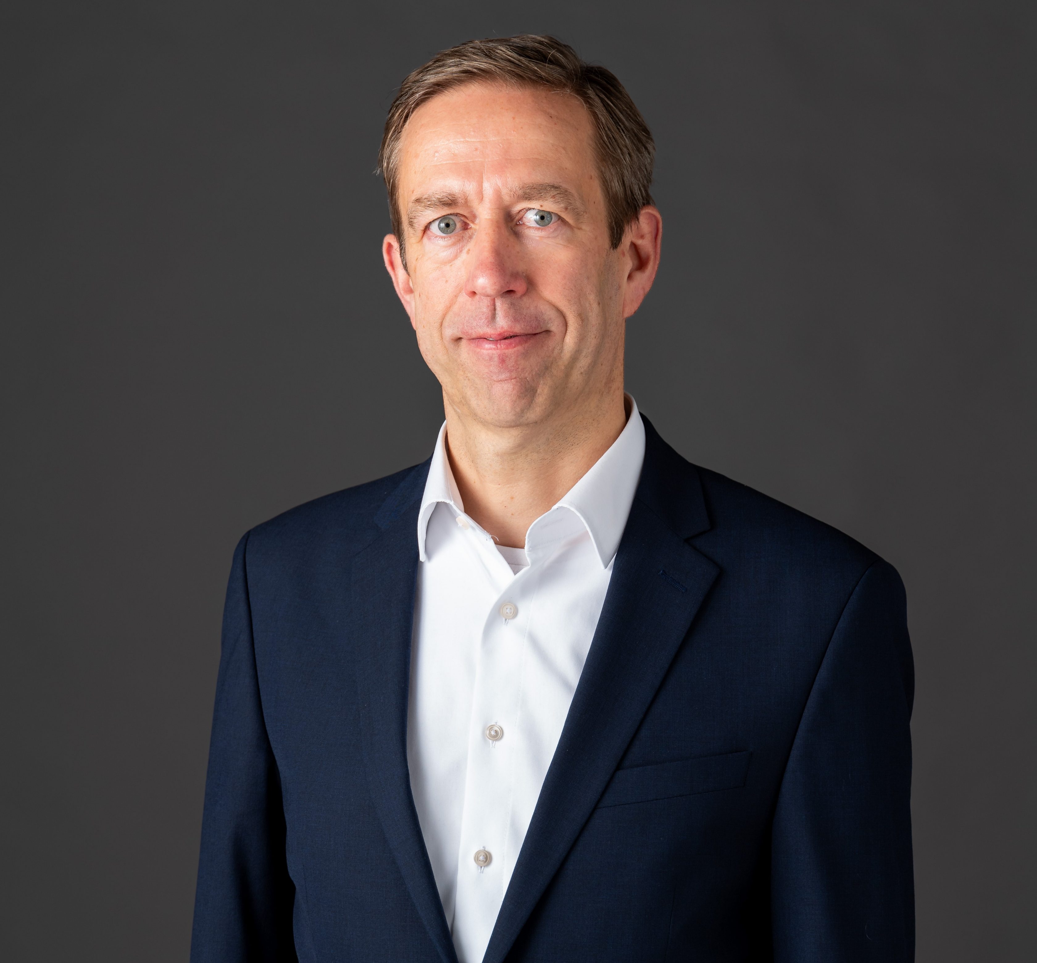 Michael Buttstedt, Chief Financial Officer von Allianz Partners: Ein Mann steht vor grauem Hintergrund. Er trägt kurze Haare, ein weißes Hemd und darüber ein Sakko.