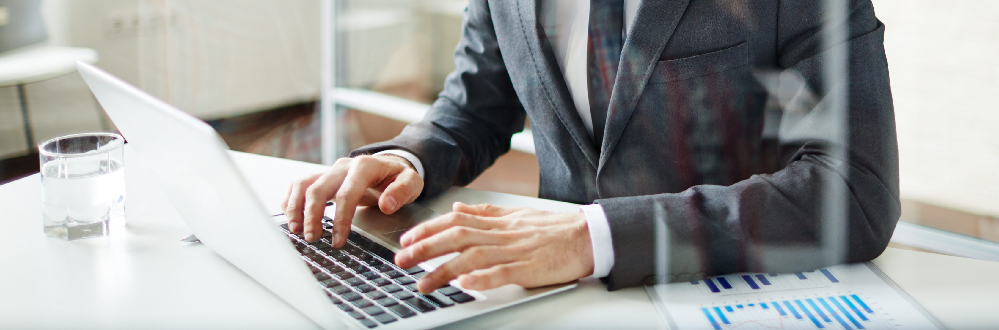 Ein Mann mit rot-braunen Haaren und Brille sitzt im Anzug am Schreibtisch und schaut auf einen geöffneten Laptop.