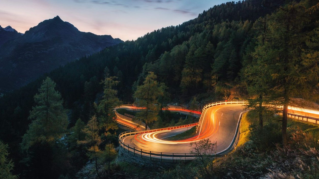 Eine kurvige Straße durch einen Wald bei Dämmerung, mit Lichtspuren von vorbeifahrenden Fahrzeugen.