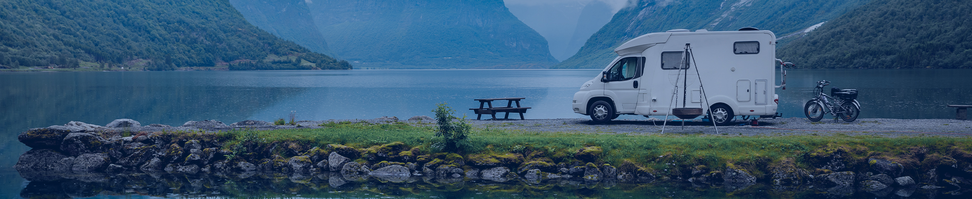 Ein Campervan steht neben einem Picknicktisch am Ufer eines Sees, umgeben von grünen Bergen und klarem Wasser.