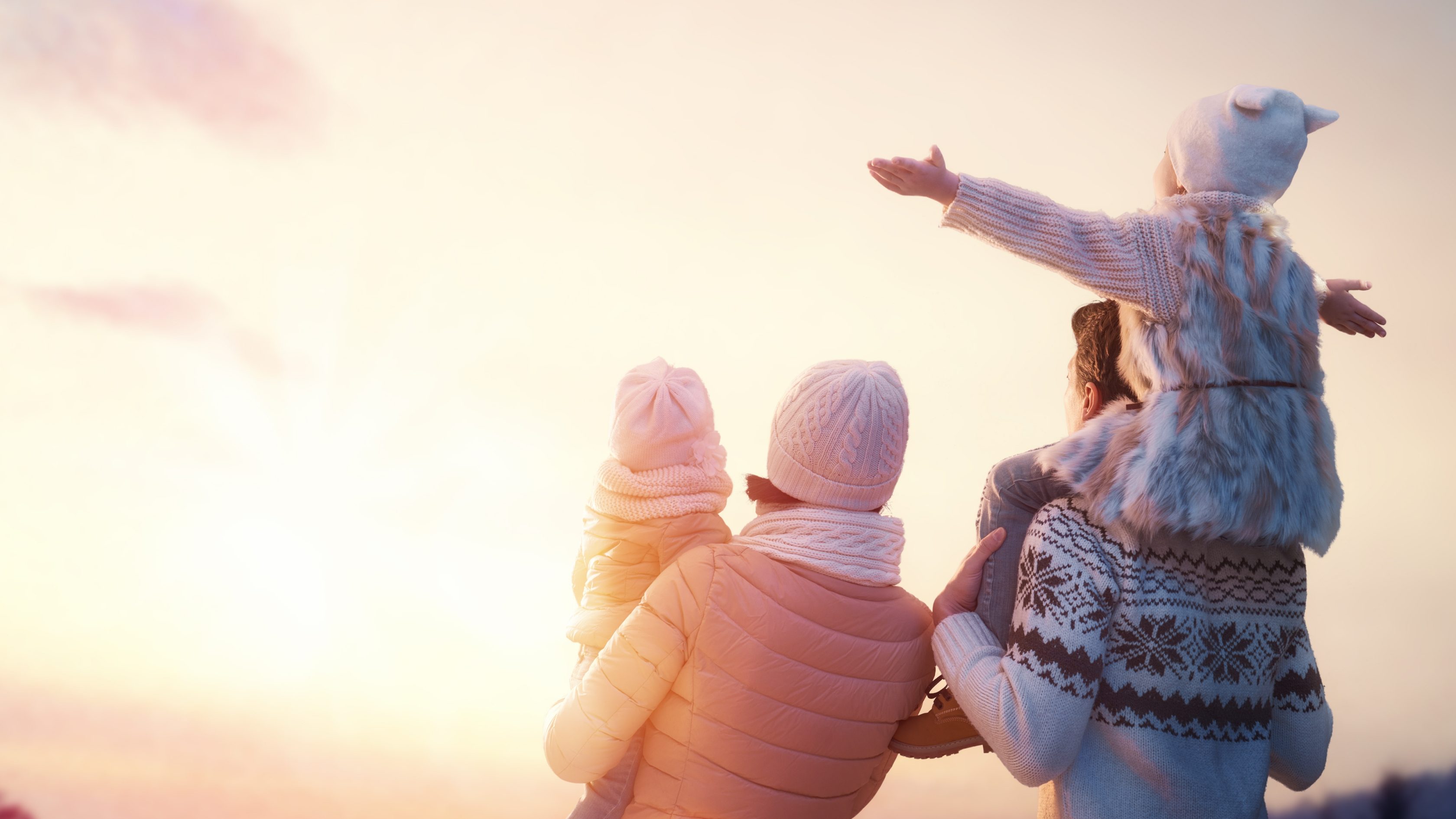 Eine vierköpfige Familie steht vor einem hellen Sonnenuntergang. Man sieht die Personen nur von hinten. Links steht die Mutter mit einem Kind auf dem Arm, rechts der Vater mit einem Kind auf den Schultern, das die Arme ausstreckt.