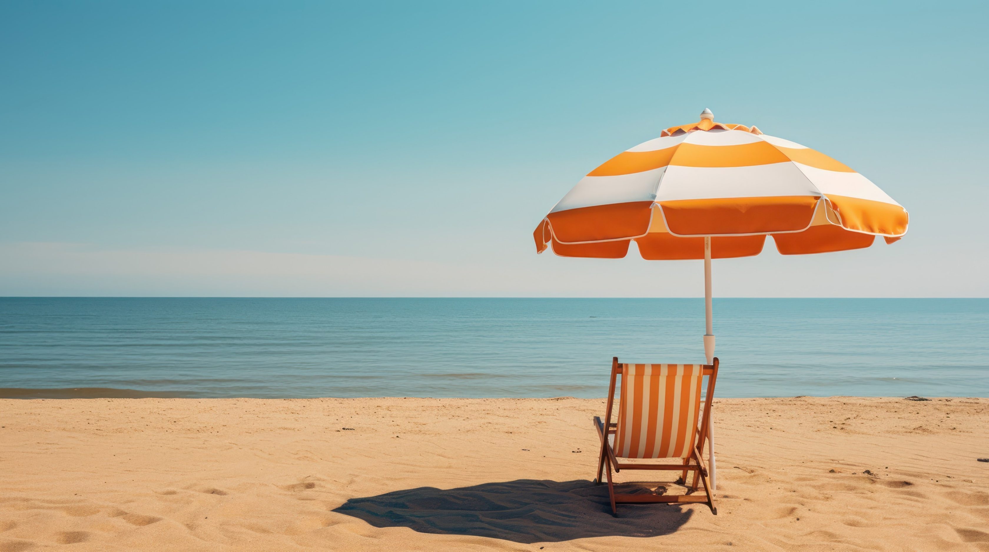 Strand im Vordergrund mit orange-weiß gestreiftem Liegestuhl und Sonnenschirm. Blaues Meer und blauer Himmel im Hintergrund.