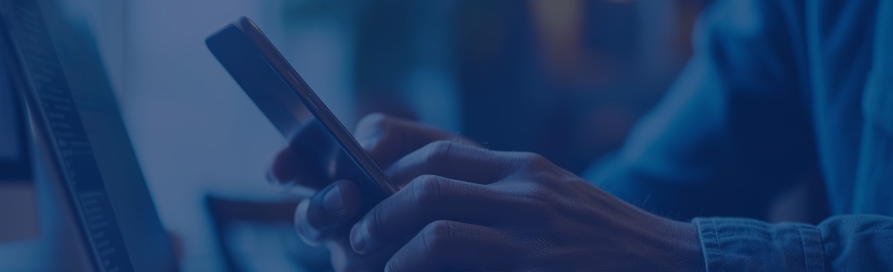 Eine Person benutzt ein Smartphone und sitzt am Tisch vor einem Laptop.