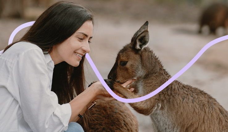 Auf dem Bild ist eine Frau mit dunklen, langen Haaren zu sehen, welche vor einem kleinen Känguru kniet und seine Pfote hält.