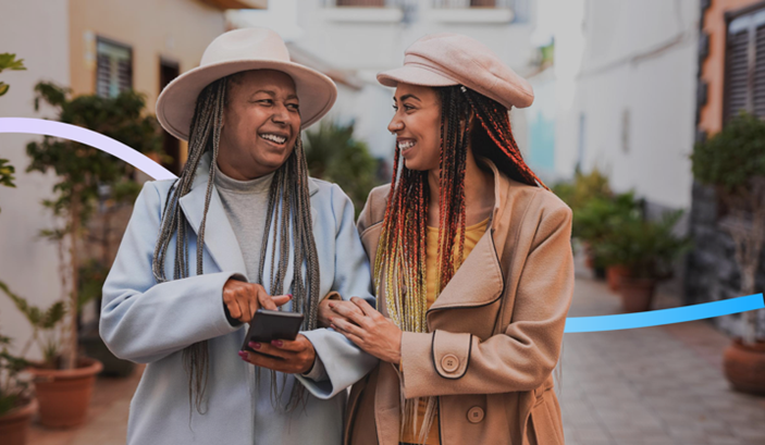 Auf dem Bild sind zwei Frau mit dunklen Braids zu sehen. Die rechte Frau ist jünger und die linke älter. Die beiden laufen eingehakt eine Straße entlang und lächeln sich an. Die linke Frau hält ein Smartphone in ihrer Hand.