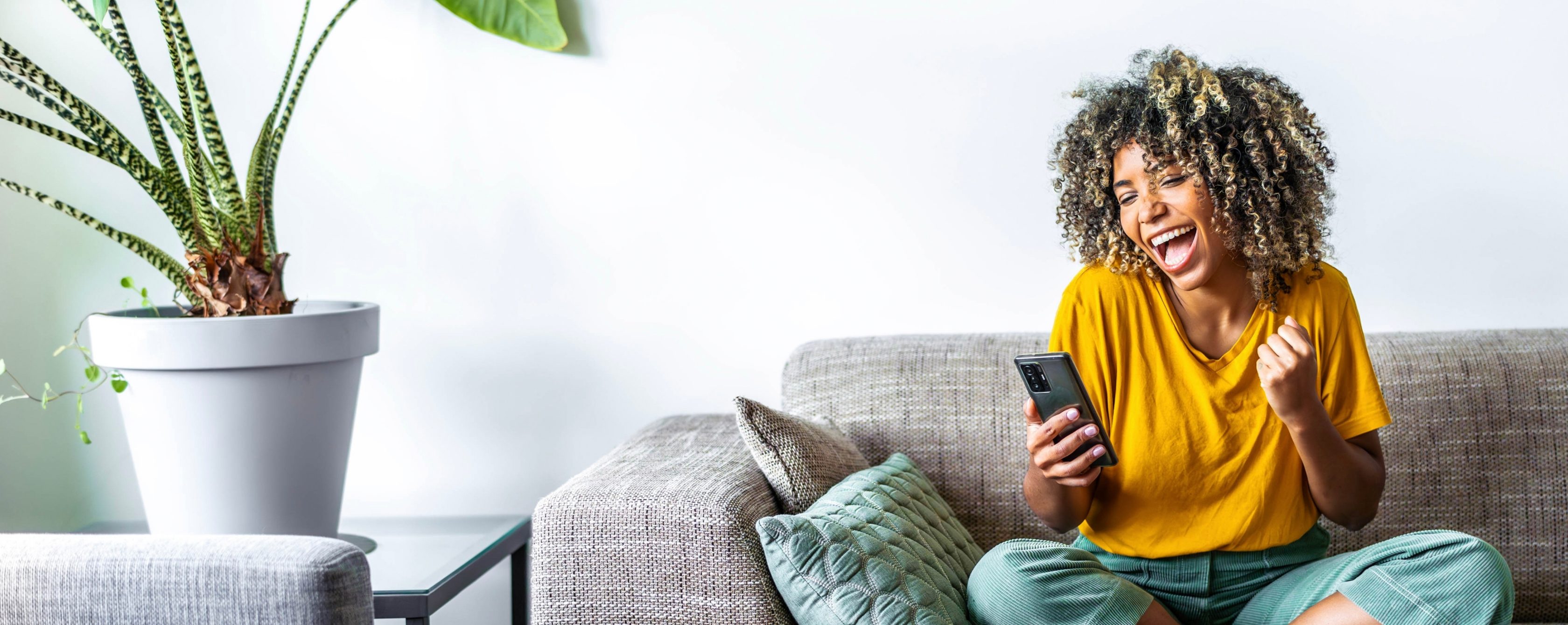 Eine Frau sitzt auf einem Sofa und schaut lachend auf ihr Smartphone. Im Hintergrund ist eine Zimmerpflanze auf einem Tisch.