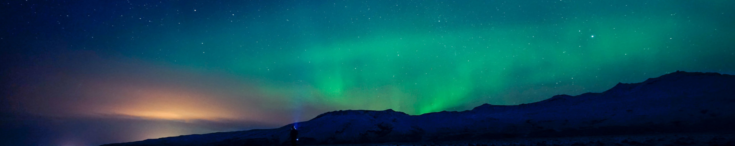 Eine Person mit Taschenlampe steht vor einer dunklen Berglandschaft, während das grüne Polarlicht den Nachthimmel erhellt.