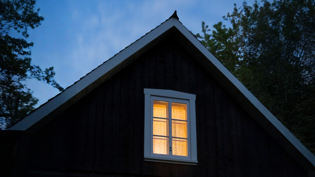 Dunkles Holzhaus mit beleuchtetem Fenster in der Dämmerung, umgeben von Bäumen.