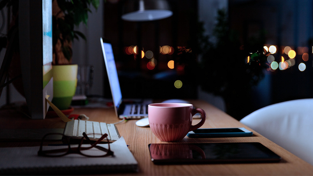 Laptop, Smartphone und rosa Kaffeetasse auf Schreibtisch im Abendlicht.