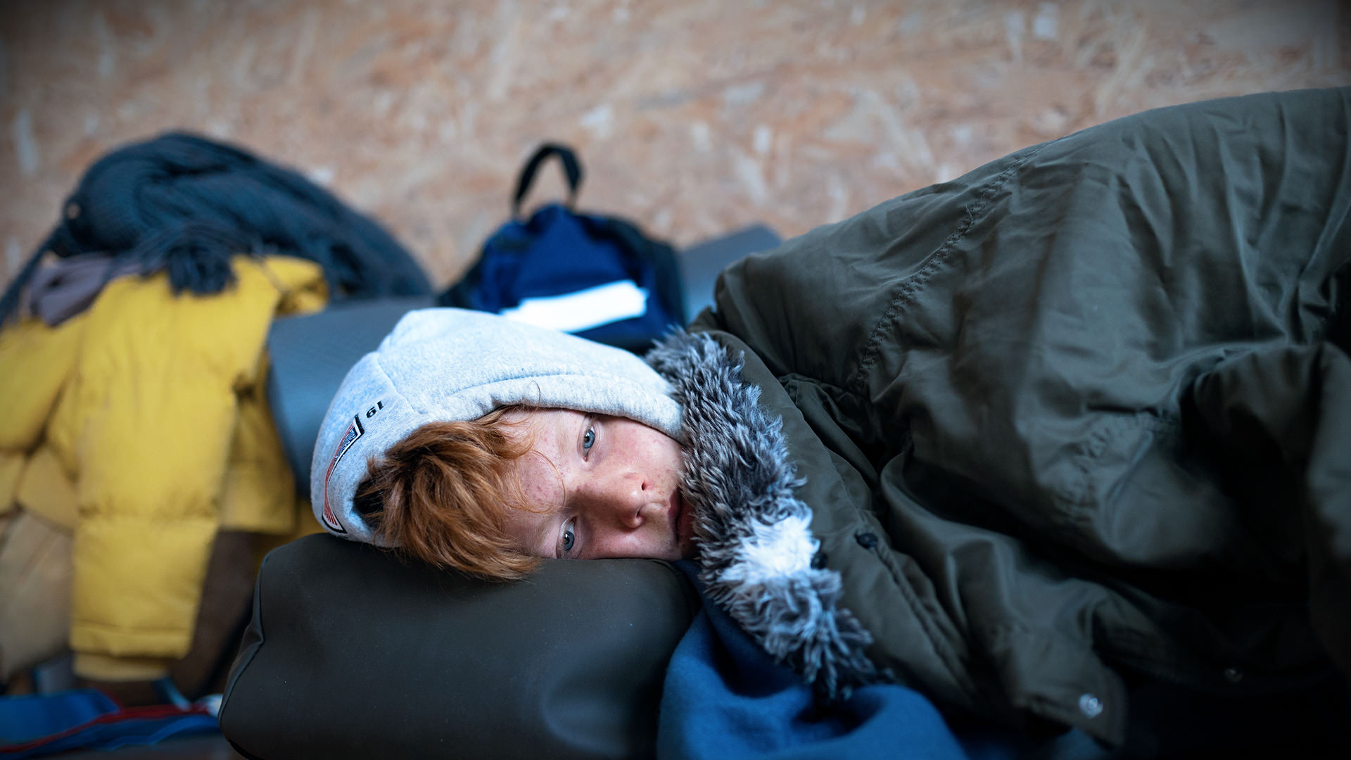 Kind mit roten Haaren liegt im Schlafsack, umgeben von Jacken und Taschen.