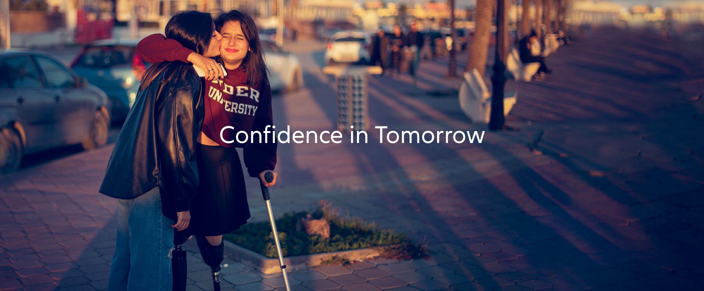 two friends, one of them with prosthetic leg, hugging each other, public place, evening sun. Text on picture: Confidence in tomorrow