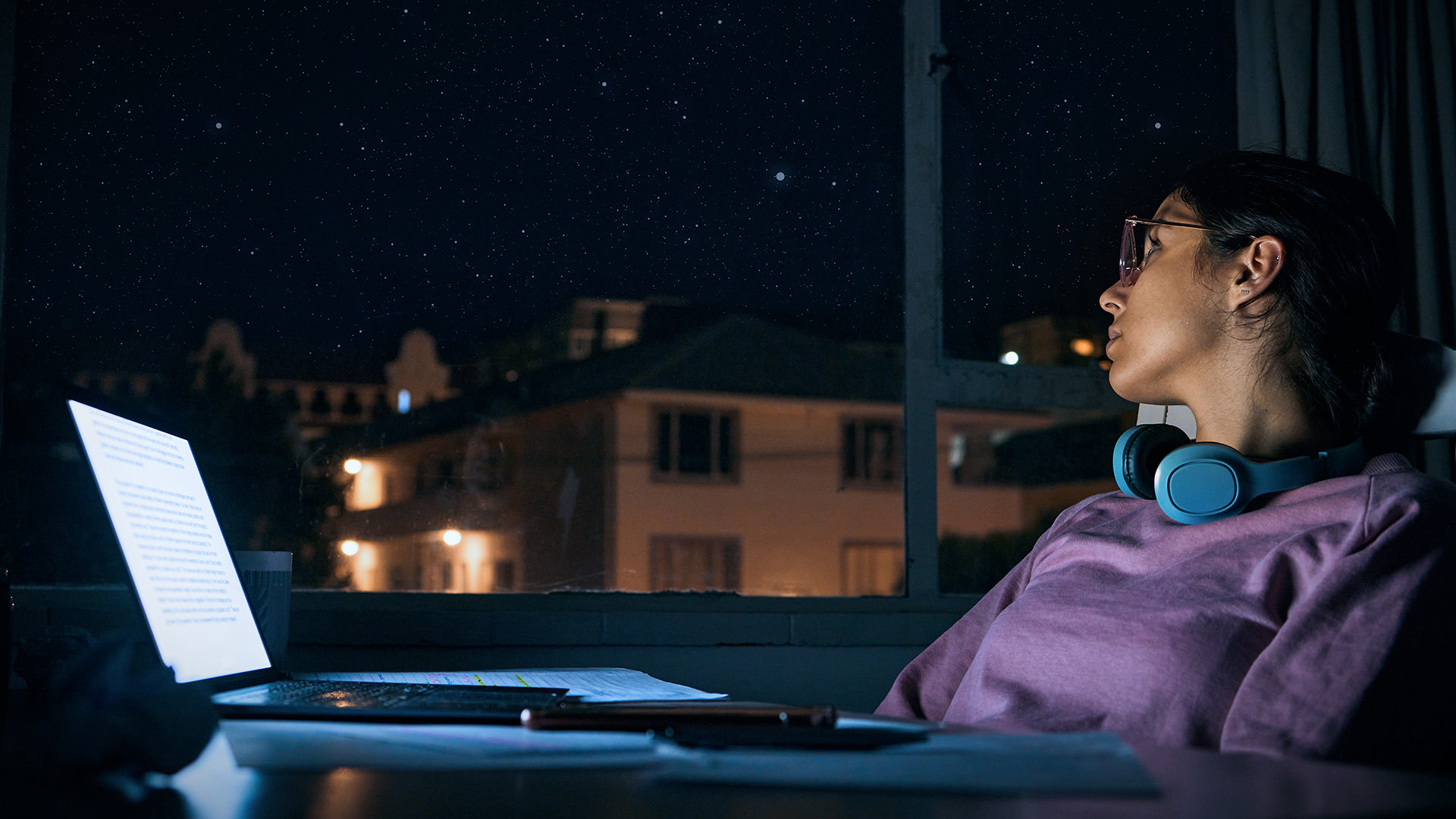 Woman with glasses and headphones looks out the window at night, laptop open on desk.