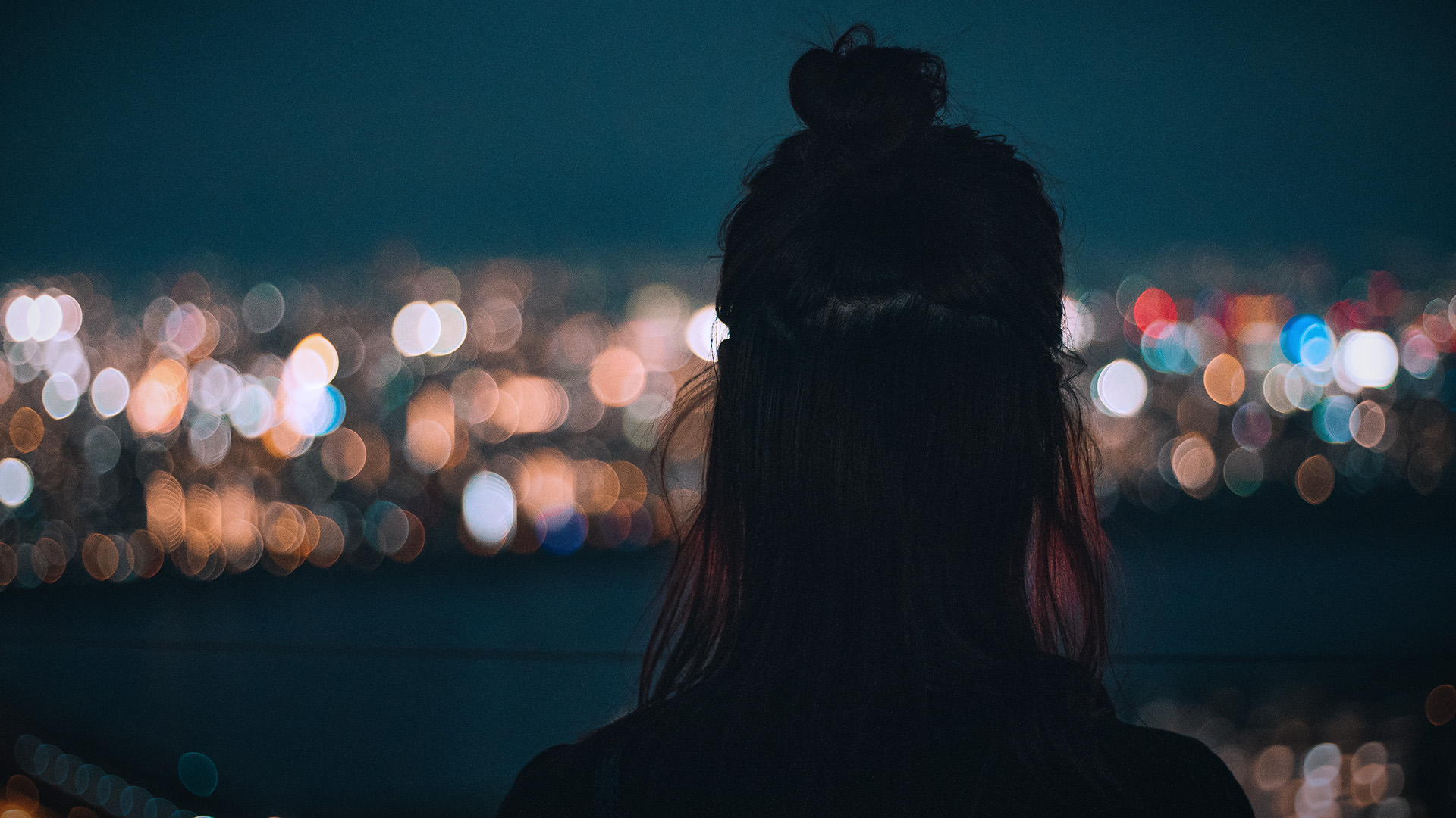 Person with dark hair seen from behind, looking over a blurred cityscape at night.
