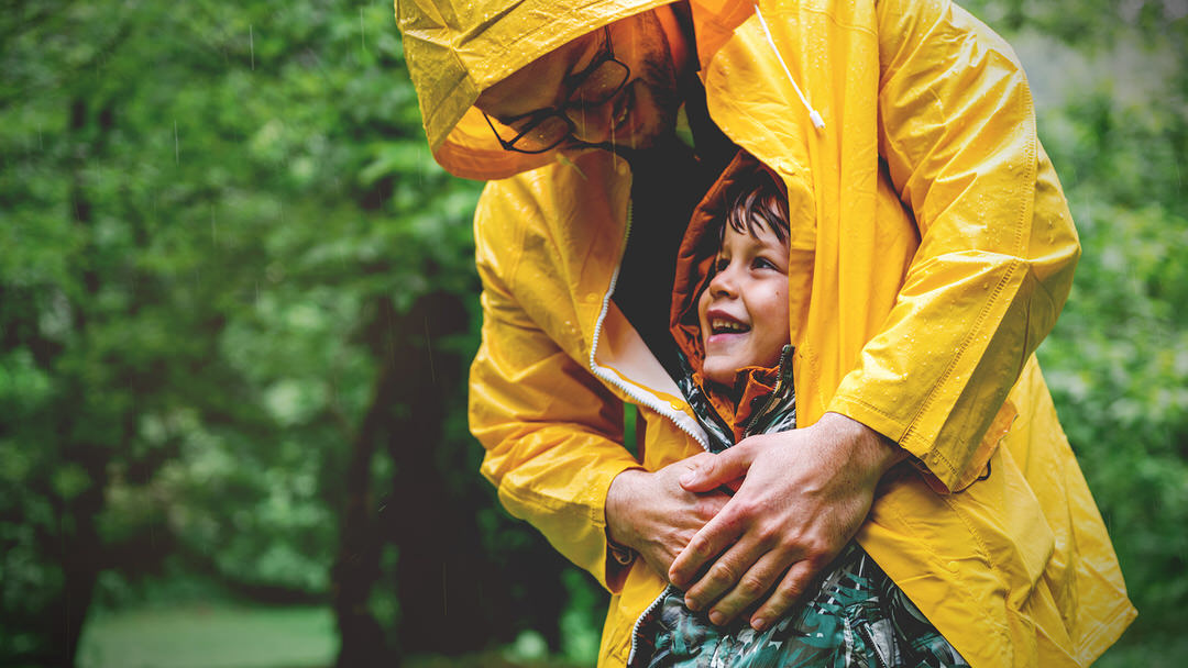 Man in yellow raincoat hugging smiling child outdoors in the rain.
