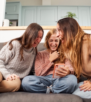 Imagen de un grupo de amigas en el sofá de casa, usando un teléfono móvil riéndose