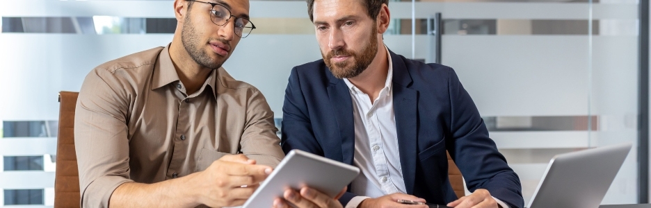 Imagen de dos empresarios revisando una tablet y un portátil mientras discuten estrategias de negocio en un entorno de oficina moderno