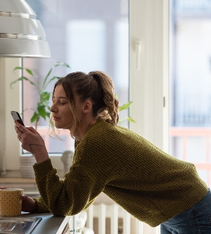 Imagen de una mujer joven sonriente usando su teléfono inteligente mientras bebe una taza de café en la cocina de casa en el concepto de la mañana mujer sonriente leyendo un mensaje telefónico