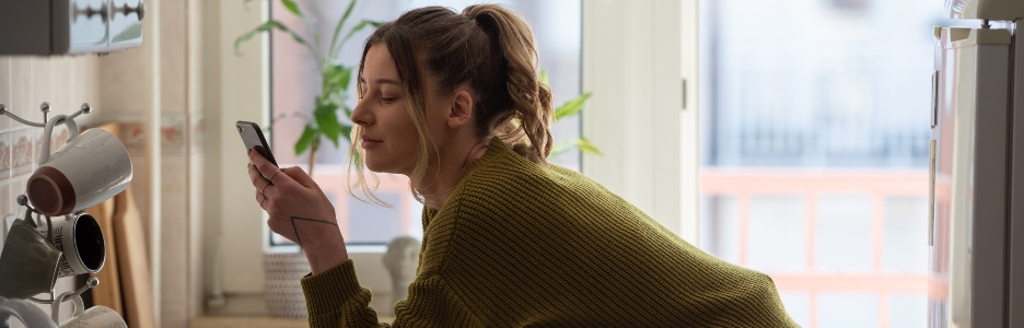 Imagen de una mujer joven sonriente usando su teléfono inteligente mientras bebe una taza de café en la cocina de casa en el concepto de la mañana mujer sonriente leyendo un mensaje telefónico