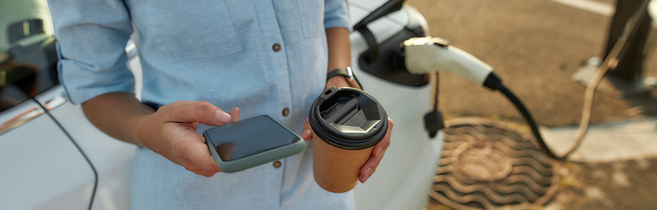 Imagen de una persona con café en mano cargando su coche eléctrico mientras navega con su teléfono móvil