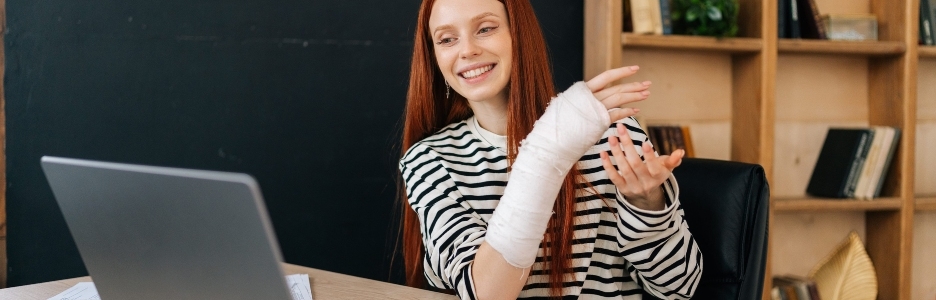 Imagen de una mujer joven y alegre con la mano derecha rota envuelta en un vendaje de yeso blanco, realizando una videollamada en línea, sonriendo y mirando a la pantalla, trabajando en remoto con un ordenador portátil, sentada en un escritorio en la habitación de su oficina en casa